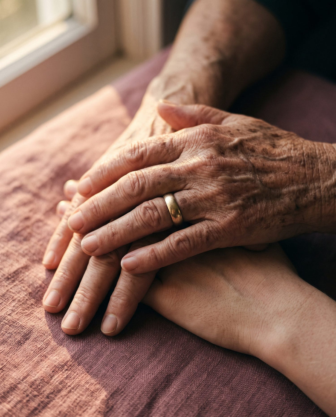 A grandmother's weathered hands with a worn wedding band resting over her daughter's hands